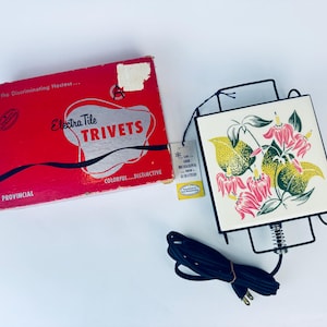 Puede incluir: Un calientaplatos vintage de baldosas eléctricas con un diseño floral. El calientaplatos es blanco con flores rosas y amarillas y hojas verdes. La caja dice "Electra Tile Trivets" y "Para la anfitriona discriminadora... Colorido... Distintivo... Provincial".