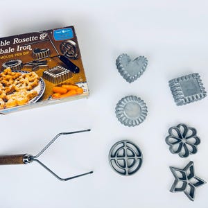 Op de afbeelding: Vintage Double Rosette & Timbal Iron set met diverse vormen. De set bevat een doos met productafbeeldingen, een handvat en diverse metalen mallen in vormen als hart, ster en bloem. Voor gebak.