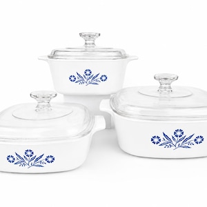 Op de afbeelding: Een set van drie witte ovenschalen met doorzichtige glazen deksels. Elke schaal heeft een blauw bloemmotief. De schalen zijn er in verschillende maten, met ronde en vierkante vormen.