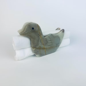 Pode incluir: Um porta-guardanapos em forma de pato cinza e castanho com um olho preto, segurando dois guardanapos brancos dobrados. O pato tem um acabamento liso e polido e uma aparência de pedra natural. Os guardanapos estão bem empilhados.