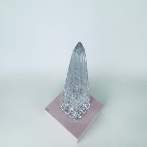 May include: A clear, faceted glass obelisk-shaped decorative object. The crystal-like structure sits on a square, light pink base. The object has a pointed top and a textured surface, reflecting light.