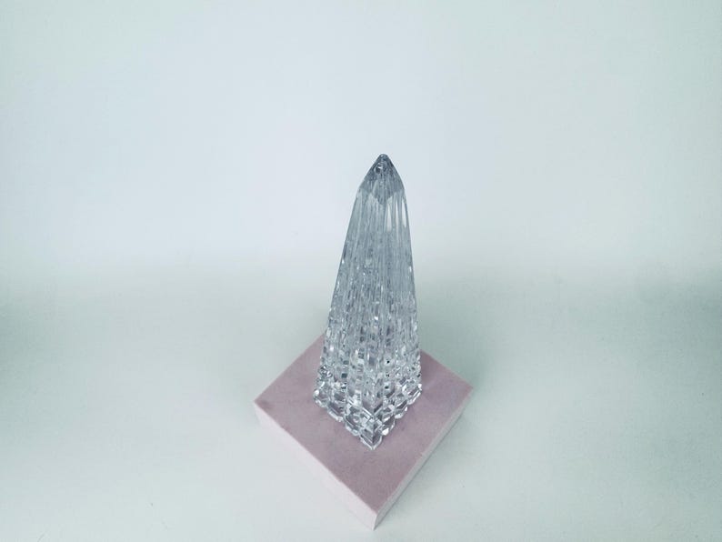 May include: A clear, faceted glass obelisk-shaped decorative object. The crystal-like structure sits on a square, light pink base. The object has a pointed top and a textured surface, reflecting light.
