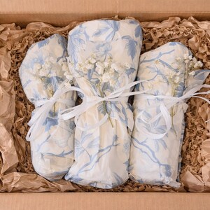 May include: Three cylindrical items wrapped in cream-colored paper with a blue floral pattern, each tied with a thin white ribbon and accented with small white dried flowers, nestled inside a cardboard box filled with crinkled brown packing paper.