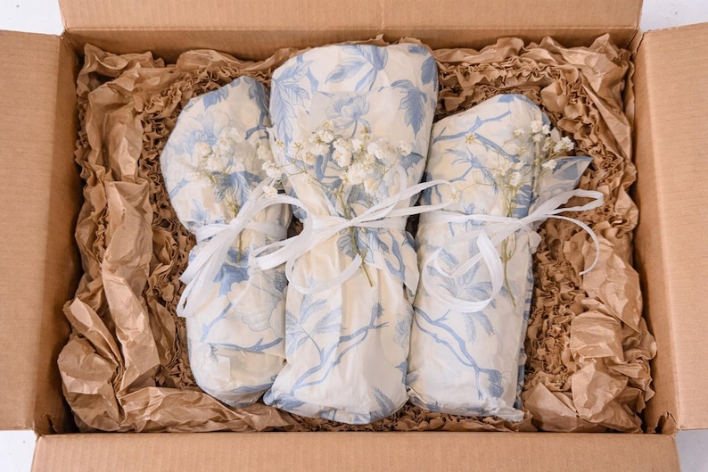 May include: Three cylindrical items wrapped in cream-colored paper with a blue floral pattern, each tied with a thin white ribbon and accented with small white dried flowers, nestled inside a cardboard box filled with crinkled brown packing paper.