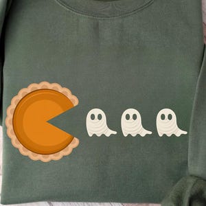 May include: An olive green sweatshirt with a pie slice design consuming three ghost figures. The pie slice is orange with a tan crust, and the ghosts are white with black eyes. The sweatshirt has long sleeves.