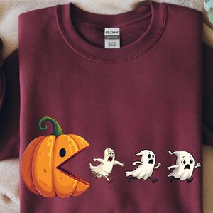 May include: A burgundy sweatshirt with a Halloween design. The graphic shows a cartoon pumpkin with an open mouth pursuing three cartoon ghosts. The sweatshirt has long sleeves and a crew neck.