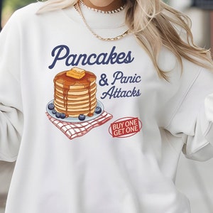 May include: White sweatshirt with a graphic of a stack of pancakes and the text "Pancakes & Panic Attacks" with a red and blue banner that says "Buy One Get One".