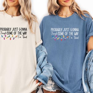 Puede incluir: Dos camisetas, una crema y otra azul, cada una con el texto "PROBABLY JUST GONNA Jingle SOME OF THE WAY I'm Tired". Una cadena de luces navide&ntilde;as de colores est&aacute; debajo del texto.