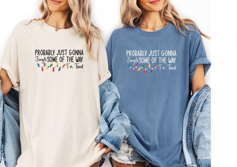 Puede incluir: Dos camisetas, una crema y otra azul, cada una con el texto "PROBABLY JUST GONNA Jingle SOME OF THE WAY I'm Tired". Una cadena de luces navide&ntilde;as de colores est&aacute; debajo del texto.