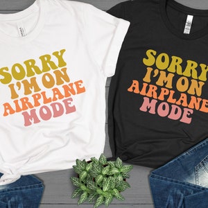 May include: Two t-shirts with the text "Sorry I'm on Airplane Mode" in a retro font with a gradient of yellow, orange, pink, and red. The t-shirts are on a gray wood background.