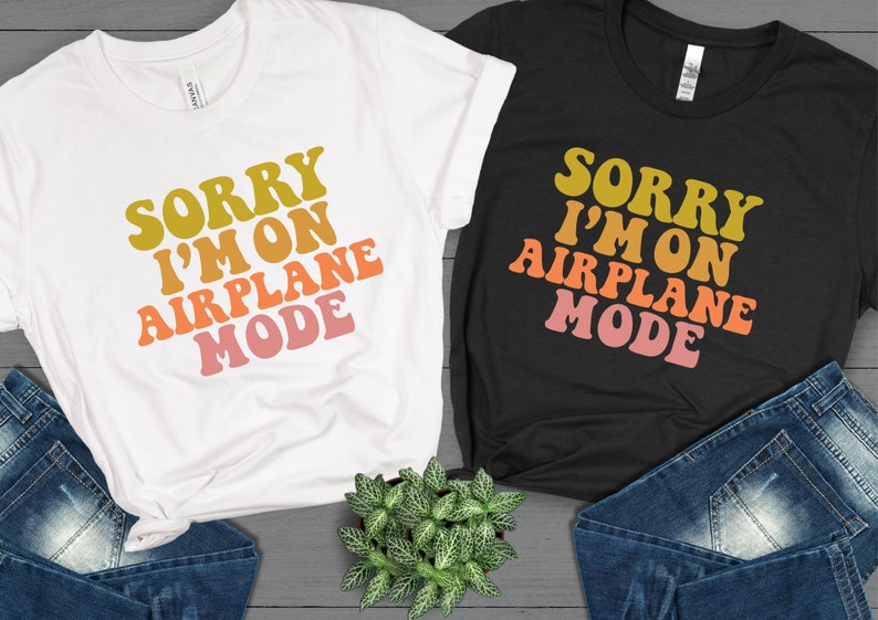 May include: Two t-shirts with the text "Sorry I'm on Airplane Mode" in a retro font with a gradient of yellow, orange, pink, and red. The t-shirts are on a gray wood background.