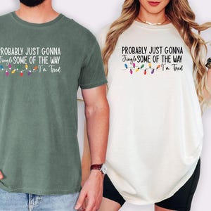 Puede incluir: Dos camisetas, una verde salvia y otra blanquecina, cada una con el texto "PROBABLY JUST GONNA Jingle SOME OF THE WAY I'm Tired" y una cadena de luces navide&ntilde;as de colores. Las camisetas son de manga corta.