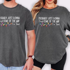 Puede incluir: Dos camisetas gris carb&oacute;n con el texto "PROBABLY JUST GONNA Jingle SOME OF THE WAY I'm Tired" en blanco, con una cadena de luces navide&ntilde;as de colores. Las camisetas son usadas por dos personas.