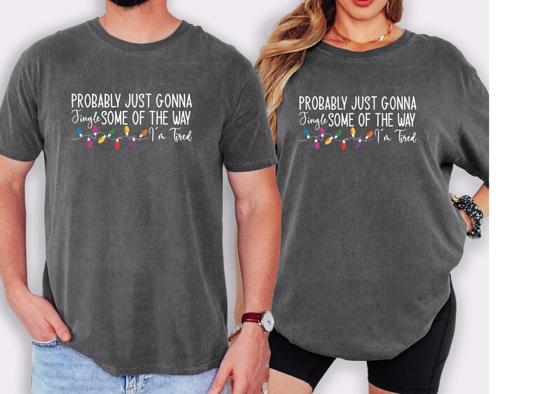Puede incluir: Dos camisetas gris carb&oacute;n con el texto "PROBABLY JUST GONNA Jingle SOME OF THE WAY I'm Tired" en blanco, con una cadena de luces navide&ntilde;as de colores. Las camisetas son usadas por dos personas.
