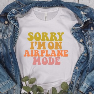 May include: White t-shirt with the text "Sorry I'm on Airplane Mode" in a retro font with yellow, orange, and pink colors.