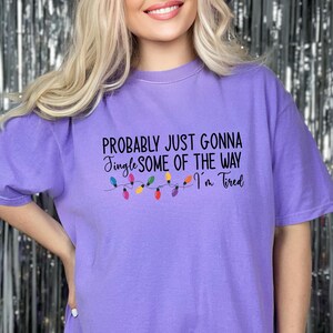 Puede incluir: Camiseta de color lila con la frase "Probably Just Gonna Jingle Some of the Way I'm Tired" en negro. Debajo del texto, una cadena de luces navide&ntilde;as de colores. La camiseta tiene mangas cortas.
