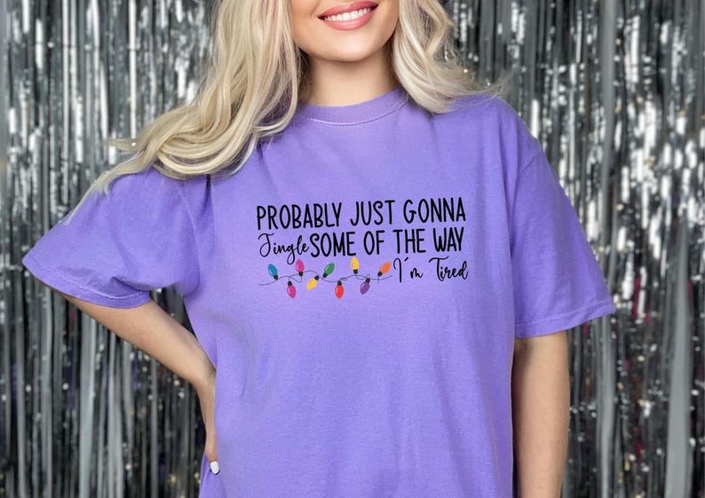 Puede incluir: Camiseta de color lila con la frase "Probably Just Gonna Jingle Some of the Way I'm Tired" en negro. Debajo del texto, una cadena de luces navide&ntilde;as de colores. La camiseta tiene mangas cortas.