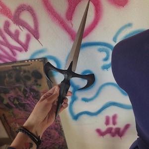 Peut inclure: Un poignard noir avec une lame argentée est tenu dans une main. Le poignard a une conception de manche unique. Le fond présente du graffiti rose et bleu sur un mur blanc.