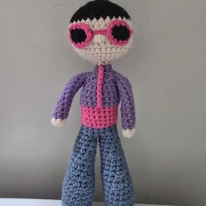 May include: A handmade crochet doll featuring a cream-colored head with black hair and pink-rimmed black sunglasses. The doll wears a purple long-sleeved top with a pink vertical stripe, a wide pink waistband, and blue trousers with white hands.