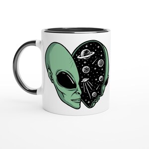 May include: White ceramic mug with a black rim and handle. The mug features a green alien head design, split to reveal a starry space scene with planets and a UFO. The alien's face is detailed with black eyes and a subtle smile.