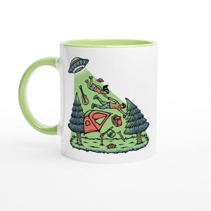 May include: White ceramic mug with a green handle and interior. The mug features a colorful illustration of a UFO beaming up two people, a tent, and camping gear. The design is set against a green and white background.