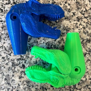 May include: Two 3D printed dinosaur heads, one blue and one green, with open mouths and visible teeth. The heads are attached to a cylindrical base.