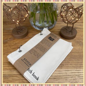 May include: A white canvas stitch book with a brown paper cover and a label that reads "STITCH  Fabric Sketchbook". The book is open and has a brown paper insert with instructions on how to use the book.