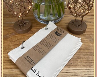 Può includere: Un libro di cucito in tela bianca con una copertina in carta marrone e un'etichetta che recita "STITCH  Fabric Sketchbook". Il libro è aperto e contiene un inserto in carta marrone con istruzioni su come utilizzare il libro.