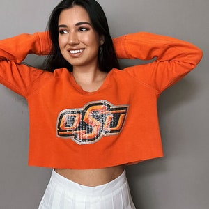 May include: A woman wearing an orange cropped sweatshirt with a sequined "OSU" logo and a white pleated skirt.
