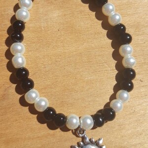 Puede incluir: Un brazalete de cuentas negras y blancas con un dije de sol plateado. El brazalete tiene un cierre plateado con un diseño de hoja.