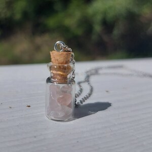 Può includere: Una collana con catena d'argento con un piccolo ciondolo a forma di bottiglia di vetro riempita di cristalli di quarzo rosa. La bottiglia ha un tappo di sughero.