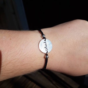 Op de afbeelding: Een armband van zwart koord met een zilveren ovale bedel waarop "I love" staat met een hartje.