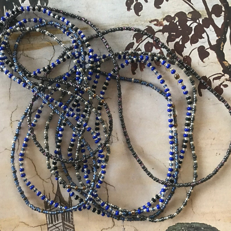 May include: A close-up of a multi-strand beaded necklace with blue, silver, and black beads. The necklace is made of small, round beads and is arranged in a loose, tangled pile.