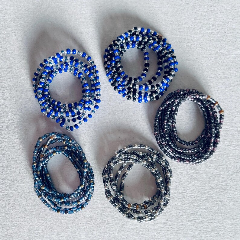 May include: Five beaded bracelets in various colors, including blue, black, and silver. The bracelets are coiled up and arranged in a circle on a white surface.