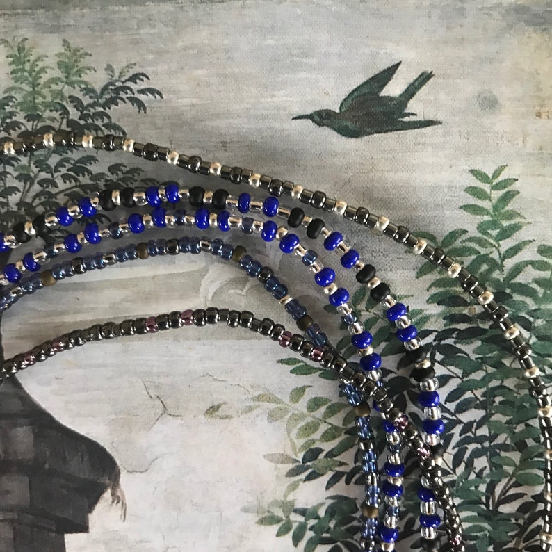 May include: A close-up of several strands of beaded jewelry. The strands are made of small, dark gray, silver, and blue beads. The beads are arranged in a pattern of alternating colors.