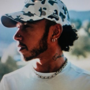 May include: A man wearing a white and black camouflage baseball cap with a white t-shirt. The t-shirt has a black Mercedes-Benz logo on the front.