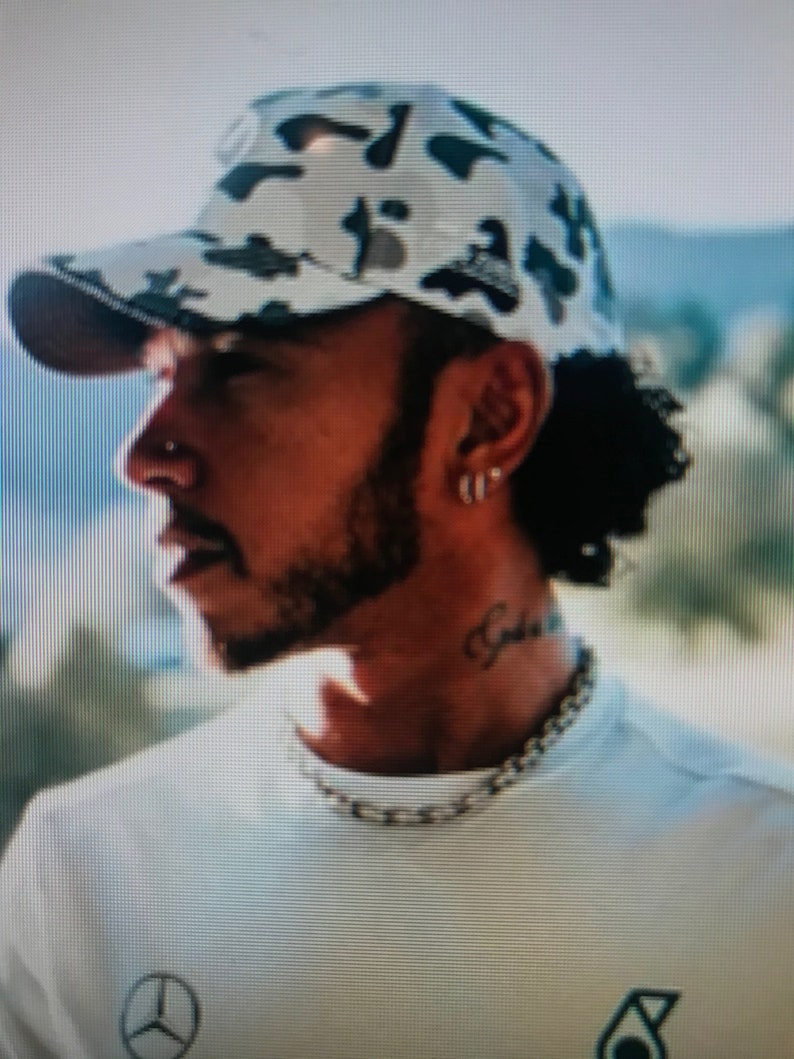 May include: A man wearing a white and black camouflage baseball cap with a white t-shirt. The t-shirt has a black Mercedes-Benz logo on the front.