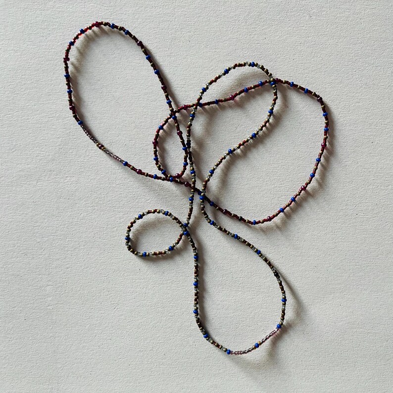 Puede incluir: Un collar largo hecho de peque&ntilde;as cuentas de color rojo oscuro, plateado y azul. El collar est&aacute; anudado y retorcido, creando un patr&oacute;n decorativo.