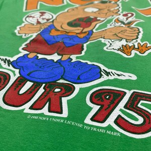 Puede incluir: Camiseta verde con un personaje de dibujos animados sosteniendo una piruleta y un p&aacute;jaro. El texto "NOFX" est&aacute; impreso en naranja y blanco sobre el personaje. El texto "TOUR 95" est&aacute; impreso en rojo y blanco debajo del personaje. El texto "&copy; 1995 NOFX UNDER LICENSE TO TRASH MARK" est&aacute; impreso en blanco debajo del personaje.