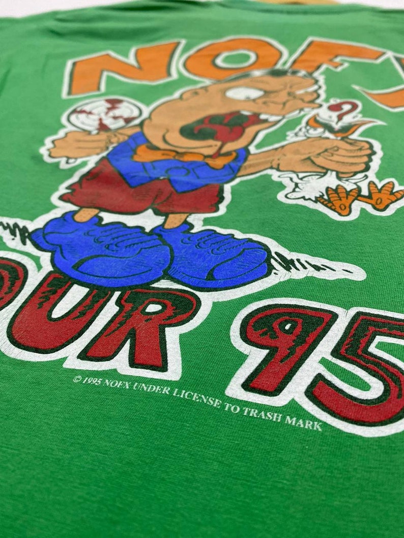 Puede incluir: Camiseta verde con un personaje de dibujos animados sosteniendo una piruleta y un p&aacute;jaro. El texto "NOFX" est&aacute; impreso en naranja y blanco sobre el personaje. El texto "TOUR 95" est&aacute; impreso en rojo y blanco debajo del personaje. El texto "&copy; 1995 NOFX UNDER LICENSE TO TRASH MARK" est&aacute; impreso en blanco debajo del personaje.