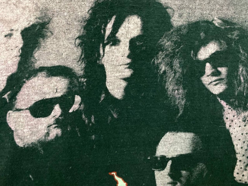 Puede incluir: Una camiseta vintage de banda en blanco y negro con una foto de cuatro personas con gafas de sol. Los miembros de la banda est&aacute;n en verde oscuro y el fondo es gris claro.