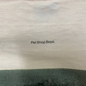 Puede incluir: Primer plano de una camiseta blanca con el texto "Pet Shop Boys." impreso en negro.