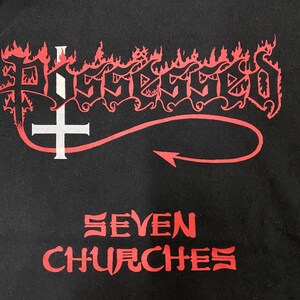 May include: Black t-shirt with a red and white graphic of the word "Possessed" in a stylized font with flames and a cross. The text "Seven Churches" is printed below the graphic.