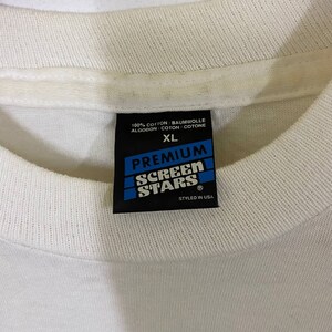 Pu&ograve; includere: Etichetta di una maglietta bianca con testo nero che recita "100% Cotone/Baumwolle/Algodon/Coton/Cotone", "XL", "Premium", "Screen Stars" e "Styled in USA".