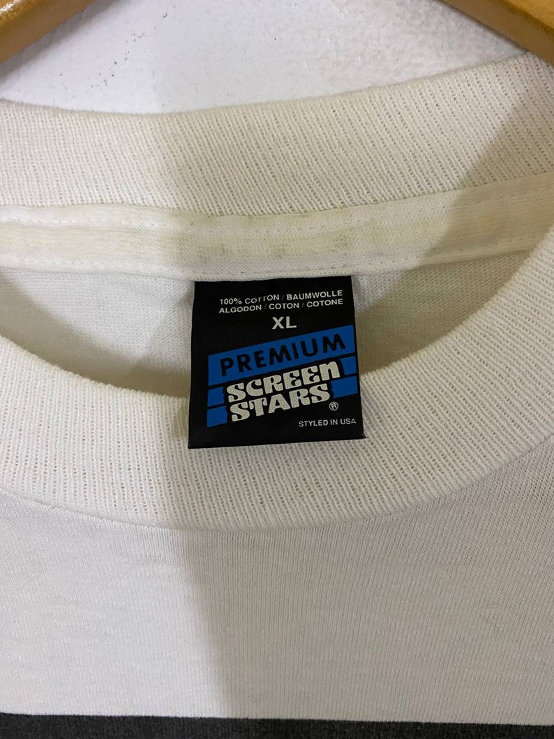 Pu&ograve; includere: Etichetta di una maglietta bianca con testo nero che recita "100% Cotone/Baumwolle/Algodon/Coton/Cotone", "XL", "Premium", "Screen Stars" e "Styled in USA".