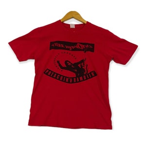 May include: Red short-sleeved t-shirt with a black and white graphic print of a person playing a guitar. The graphic includes the text "Three Hypnotics" and "Preachin' & Ramblin'"