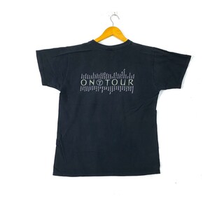 Może przedstawiać: Czarny T-shirt z kr&oacute;tkim rękawem z nadrukiem graficznym tekstu "ON TOUR" w kolorze białym i zielonym. Grafika zawiera r&oacute;wnież małe k&oacute;łko z literą "T" w środku.
