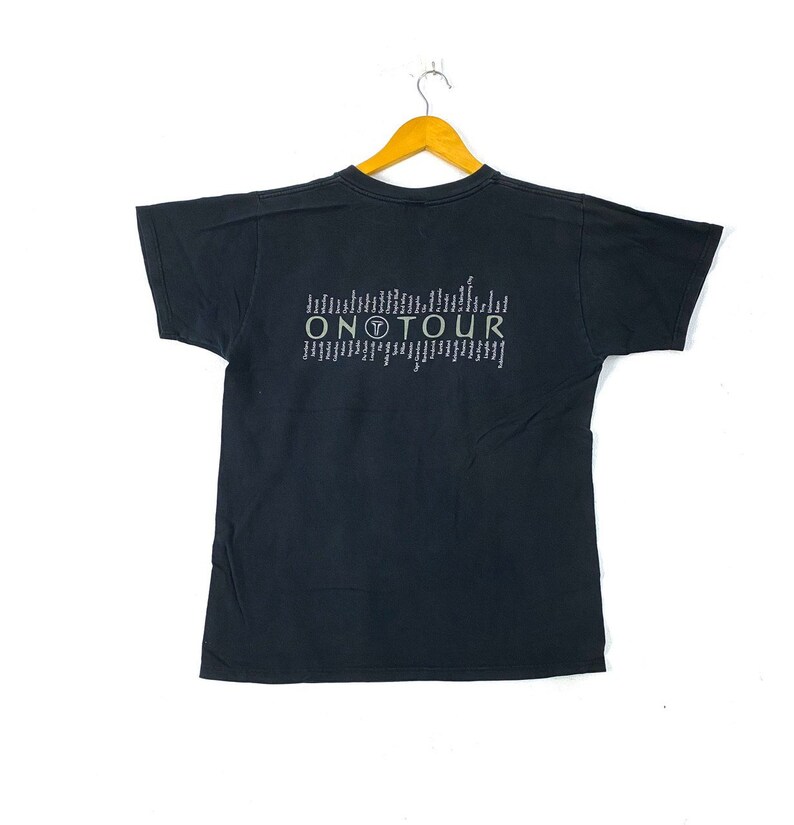 Może przedstawiać: Czarny T-shirt z kr&oacute;tkim rękawem z nadrukiem graficznym tekstu "ON TOUR" w kolorze białym i zielonym. Grafika zawiera r&oacute;wnież małe k&oacute;łko z literą "T" w środku.