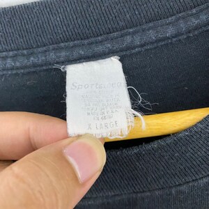 Puede incluir: Una etiqueta blanca con texto negro que dice "Sporty's" y "Made in USA" adjunta a una camiseta negra. La etiqueta tambi&eacute;n dice "All Sizes" y "Machine Wash Cold".