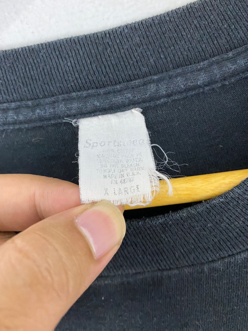 Puede incluir: Una etiqueta blanca con texto negro que dice "Sporty's" y "Made in USA" adjunta a una camiseta negra. La etiqueta tambi&eacute;n dice "All Sizes" y "Machine Wash Cold".